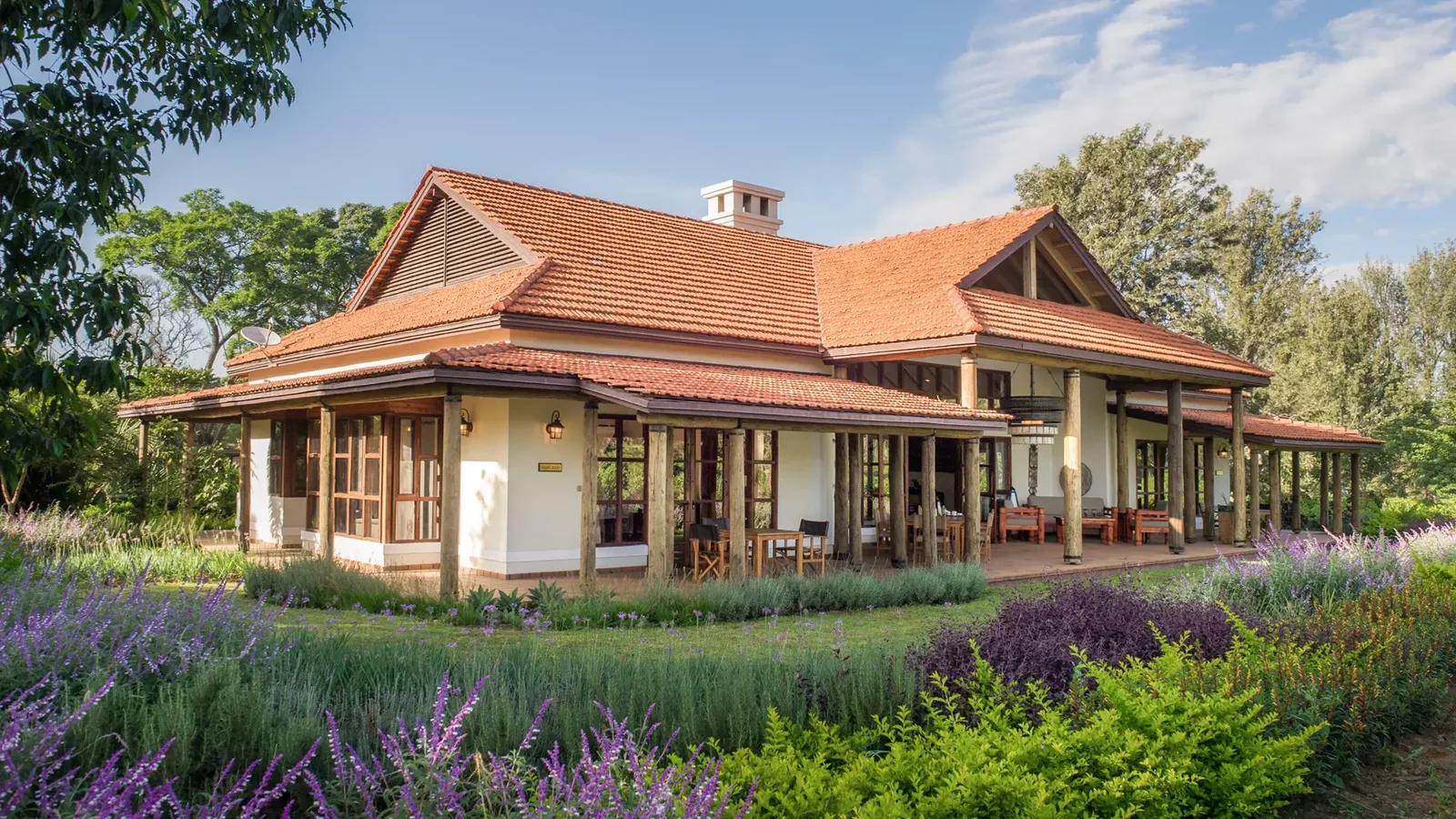Charming lodge with a terracotta roof surrounded by lush gardens and flowering plants under a clear blue sky.