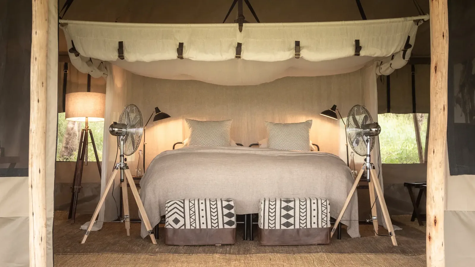 Luxury safari tent interior featuring a king-sized bed, soft neutral fabrics, tripod lamps, and rustic wooden details.